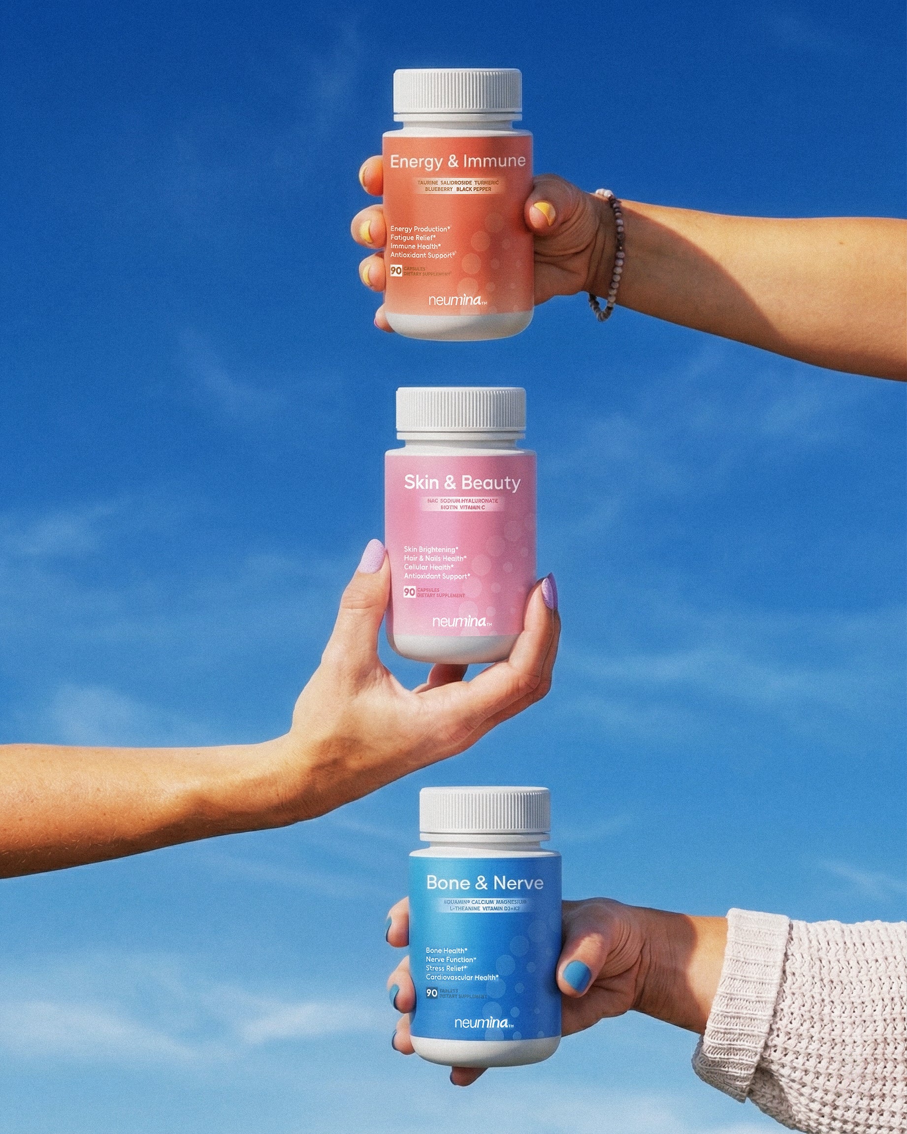 A promotional lifestyle photograph featuring three Neumina women's wellness supplements held against a clear blue sky. From top to bottom: 1) Energy & Immune (Orange) for vitality. 2) Skin & Beauty (Pink) featuring NAC, Biotin, and Hyaluronic Acid for brightening and hydration. 3) Bone & Nerve (Blue) for structural and neurological support. Each bottle contains 90 capsules. This visual represents a holistic daily nutrition stack designed for the modern woman to support beauty, immunity, and strength.
